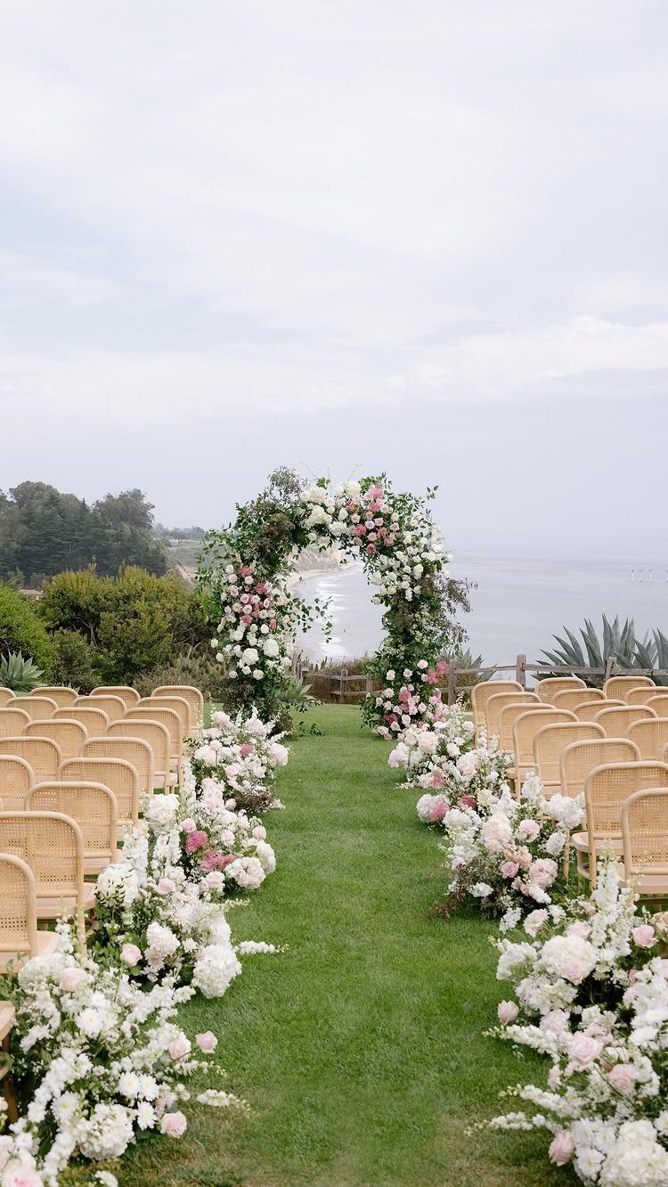 Floral wedding arch with pink and white flowers overlooking the ocean on grass.