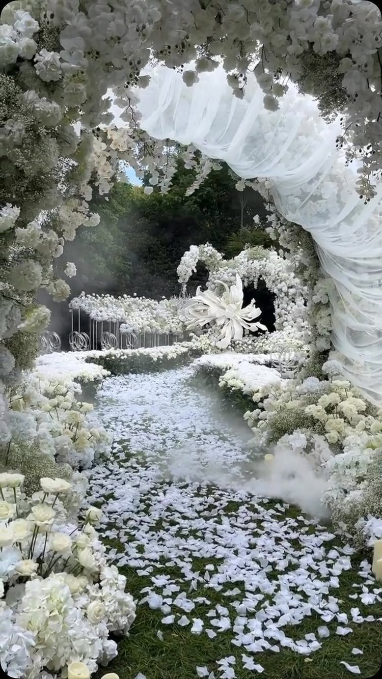 White floral arches with draped fabric over a petal-covered grass wedding aisle.