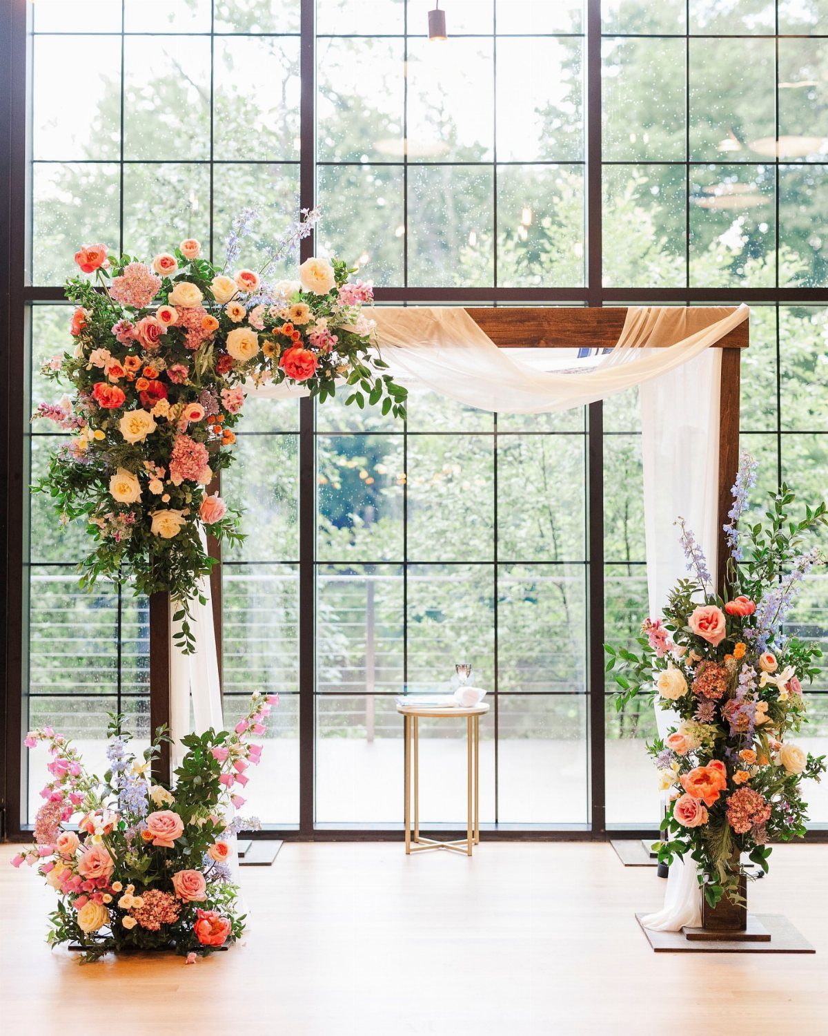 Wooden wedding arch decorated with colorful floral arrangements and white fabric draping.