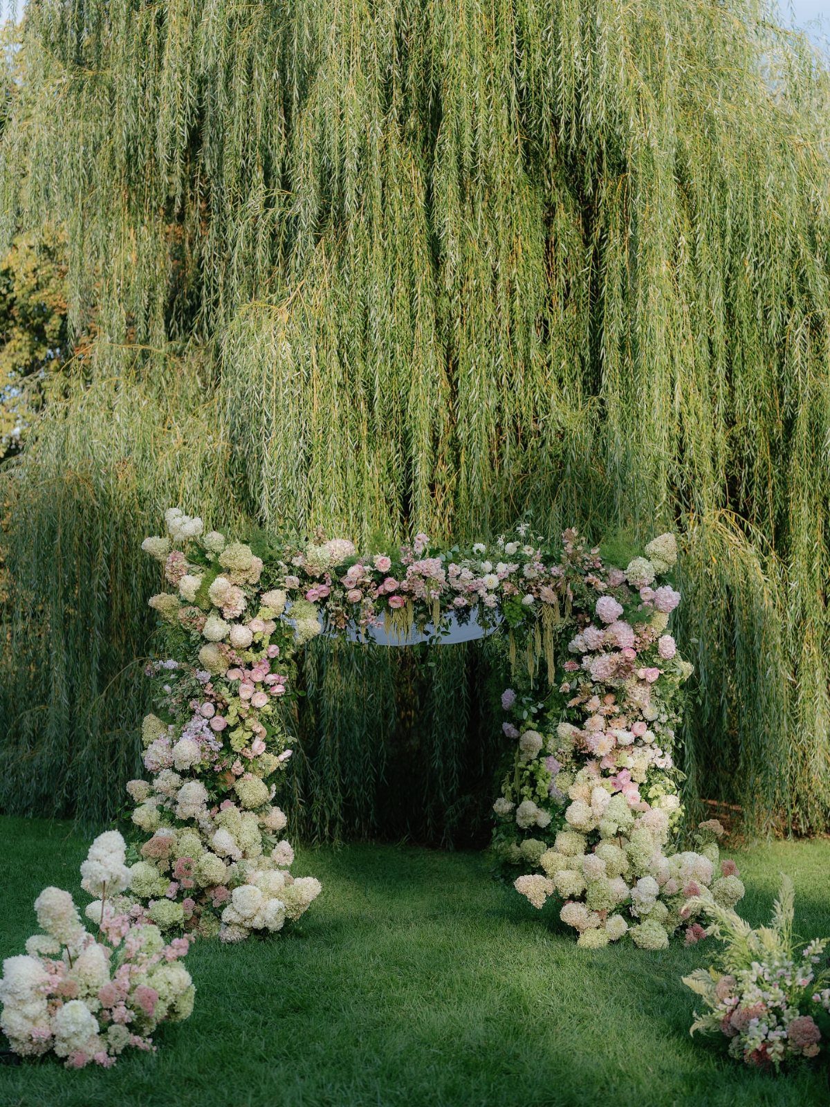Floral wedding arch with white and pink roses before a weeping willow tree.