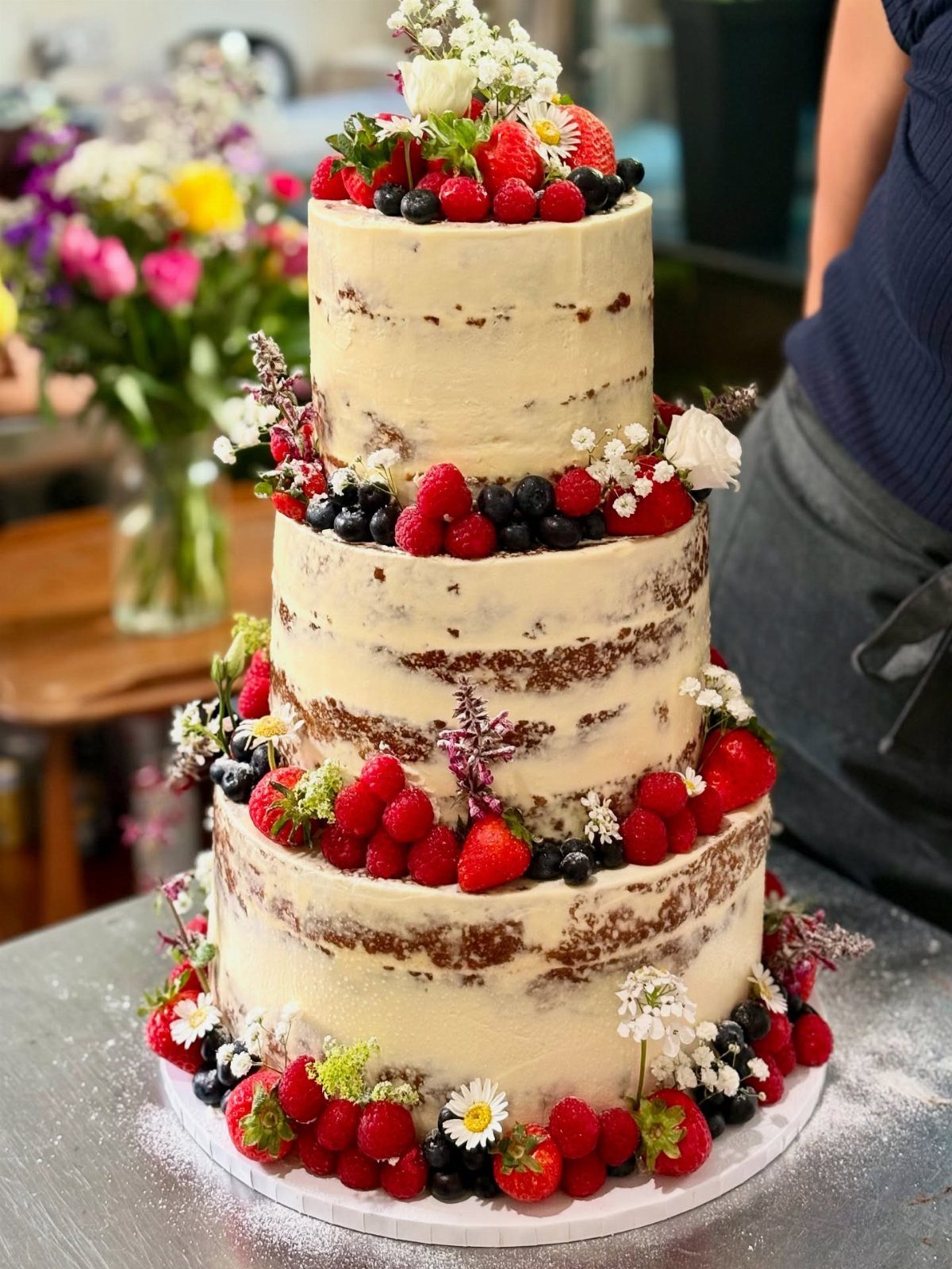 Three-tiered semi-naked cake decorated with fresh berries and small white flowers.