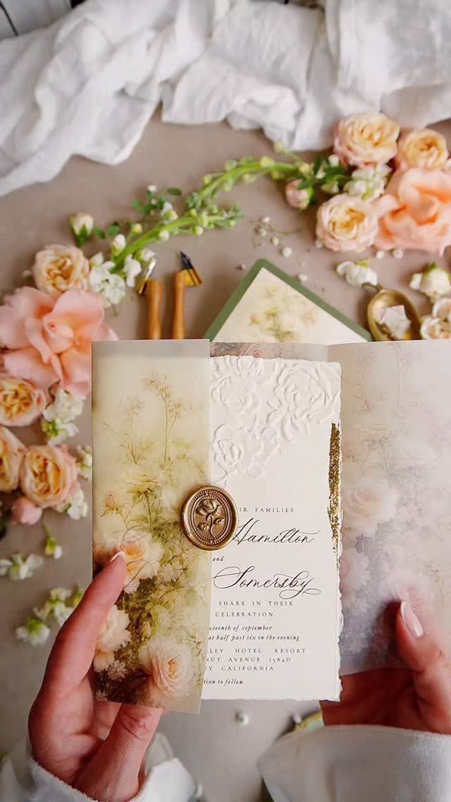 Floral wedding invitation with a gold wax seal held by two hands.