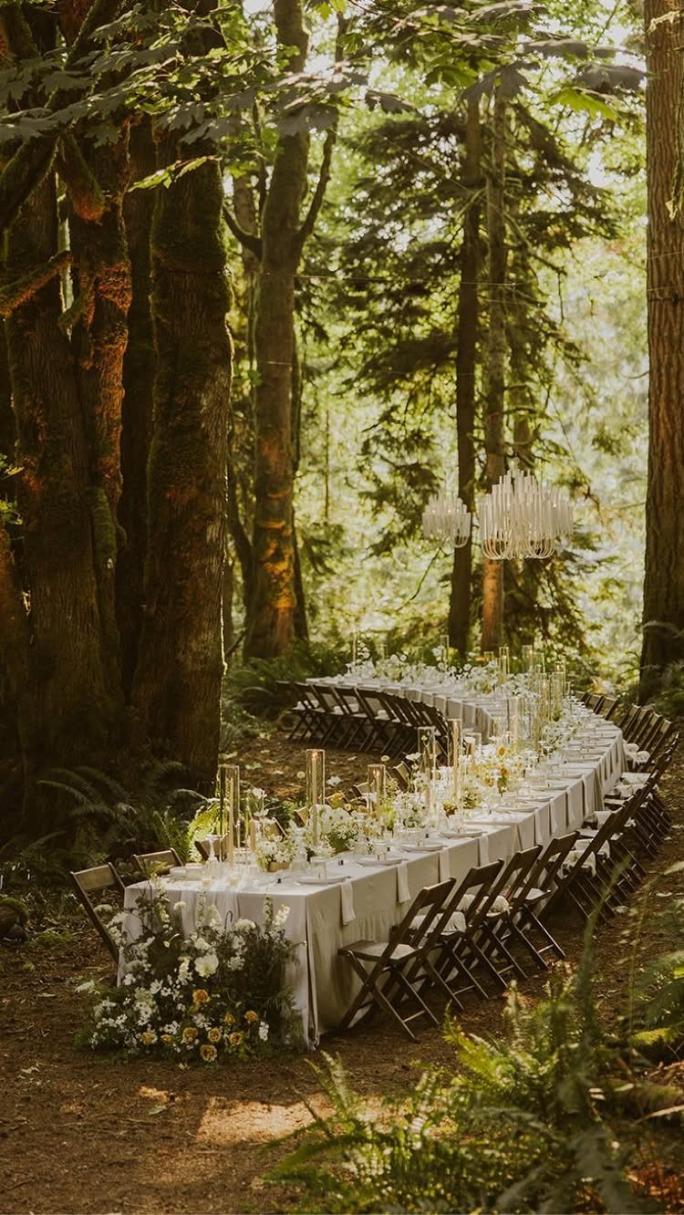 Long curved white dining table with floral arrangements in a sunlit forest setting