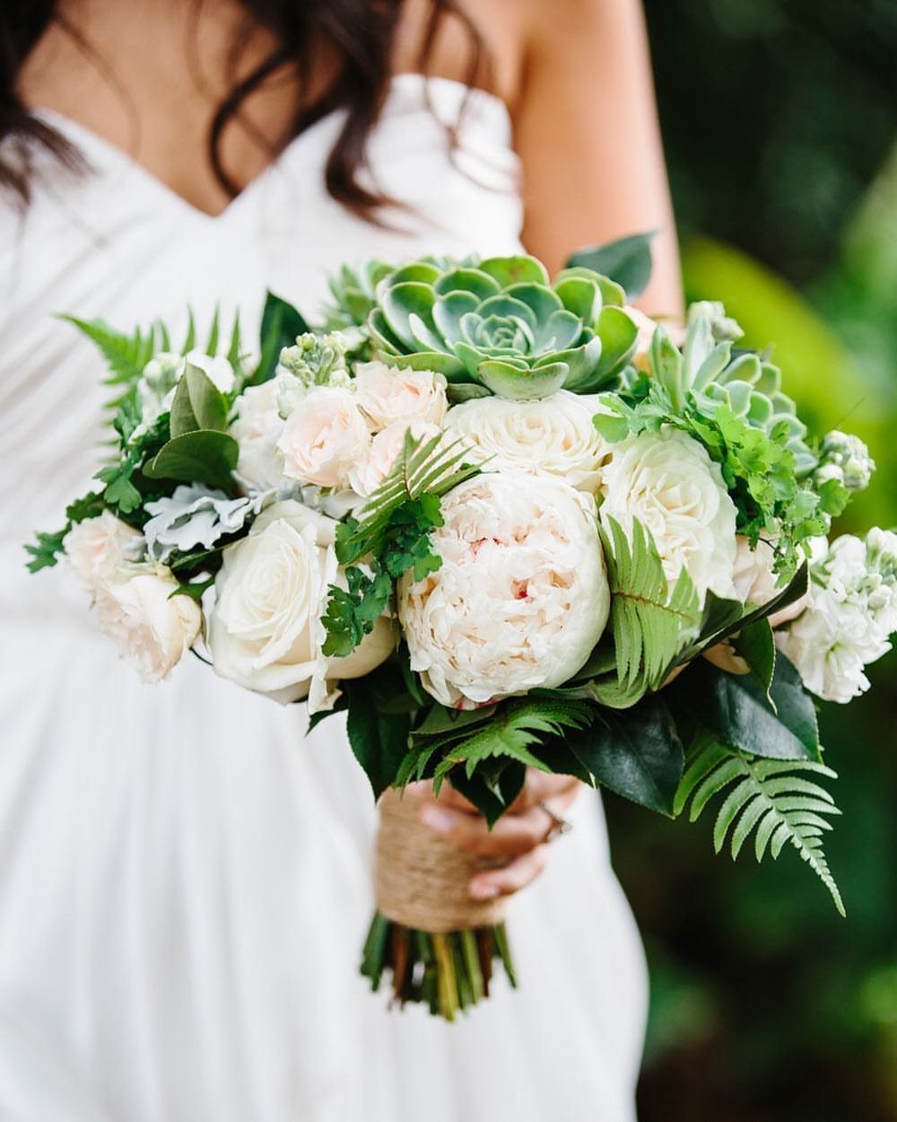 Wedding bouquet with white roses, succulents, and green fern leaves held by bride