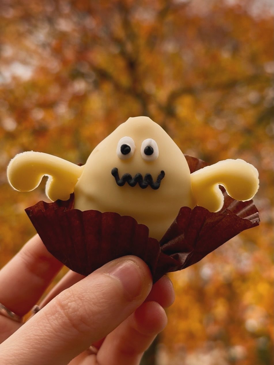 Ghost-shaped cake pop in a brown liner held against autumn foliage background