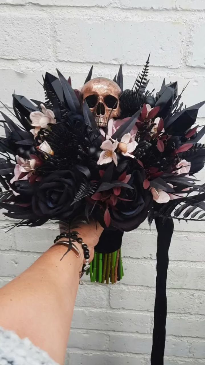 Gothic wedding bouquet with black roses and a gold skull against white brick.