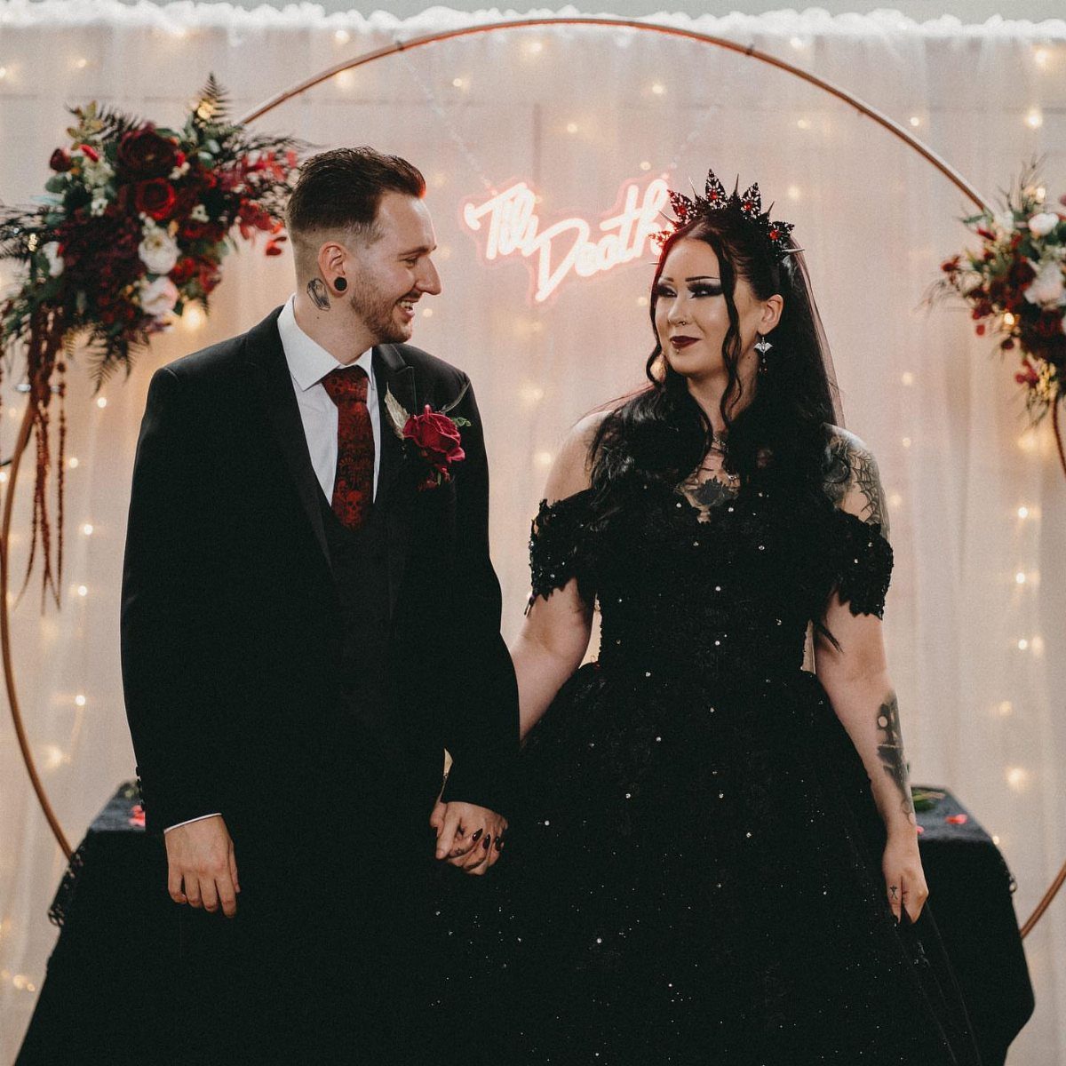 Gothic couple in black wedding attire holding hands before a circular floral arch.