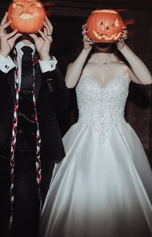 Bride and groom holding carved pumpkins over their faces in wedding attire