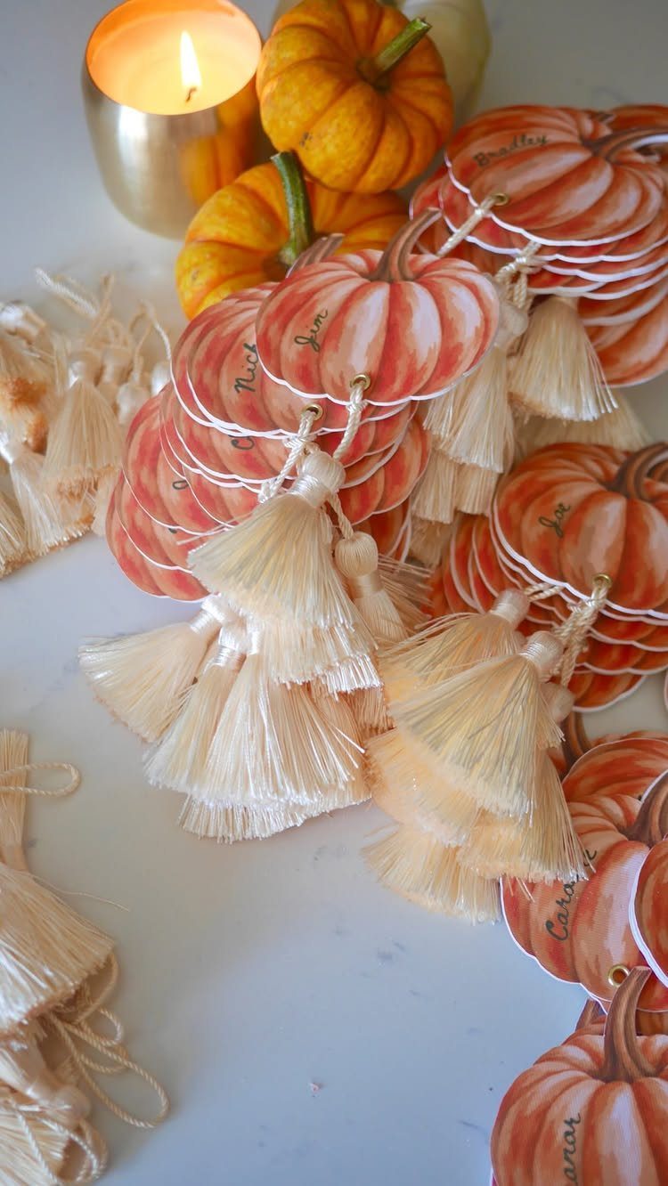 Pumpkin-shaped place cards with cream tassels and mini pumpkins on a white table.