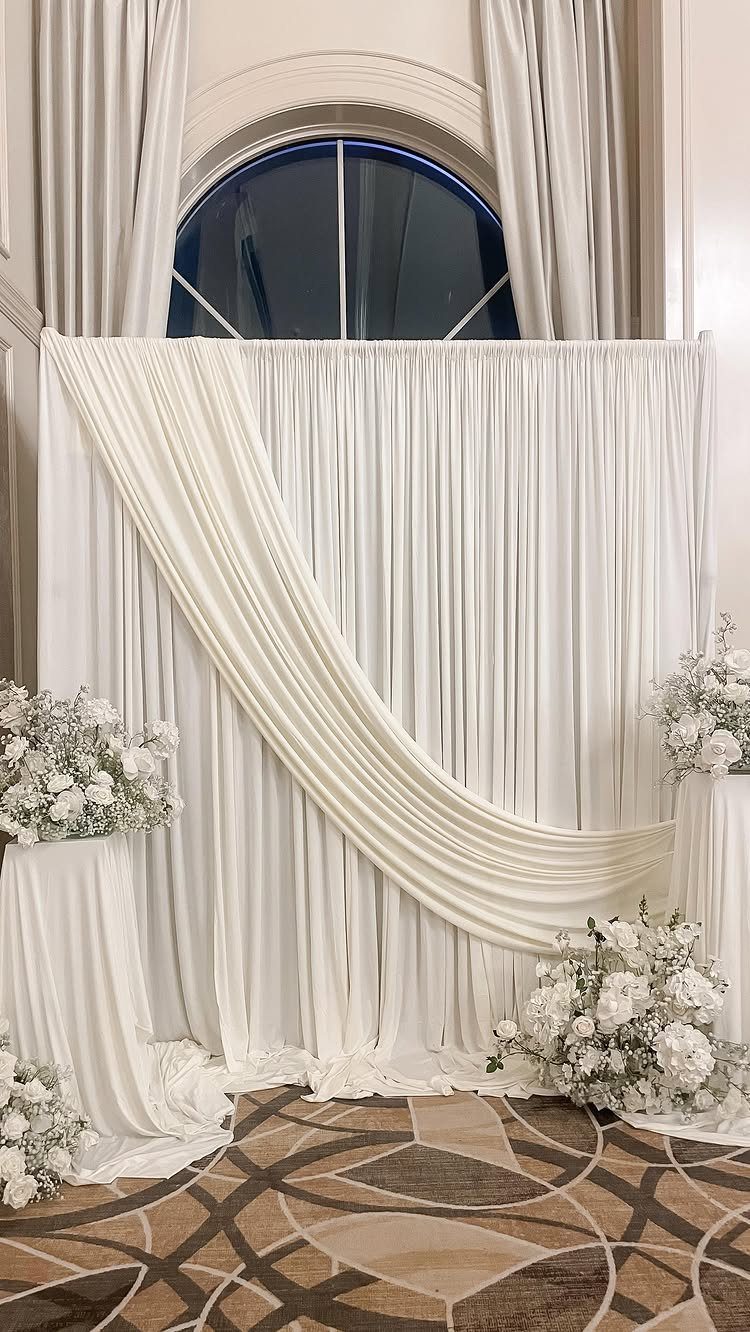 White draped fabric photo backdrop with white floral arrangements on a patterned carpet.