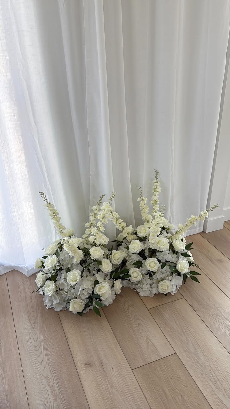 White floral arrangements with roses on a wooden floor against white curtains.