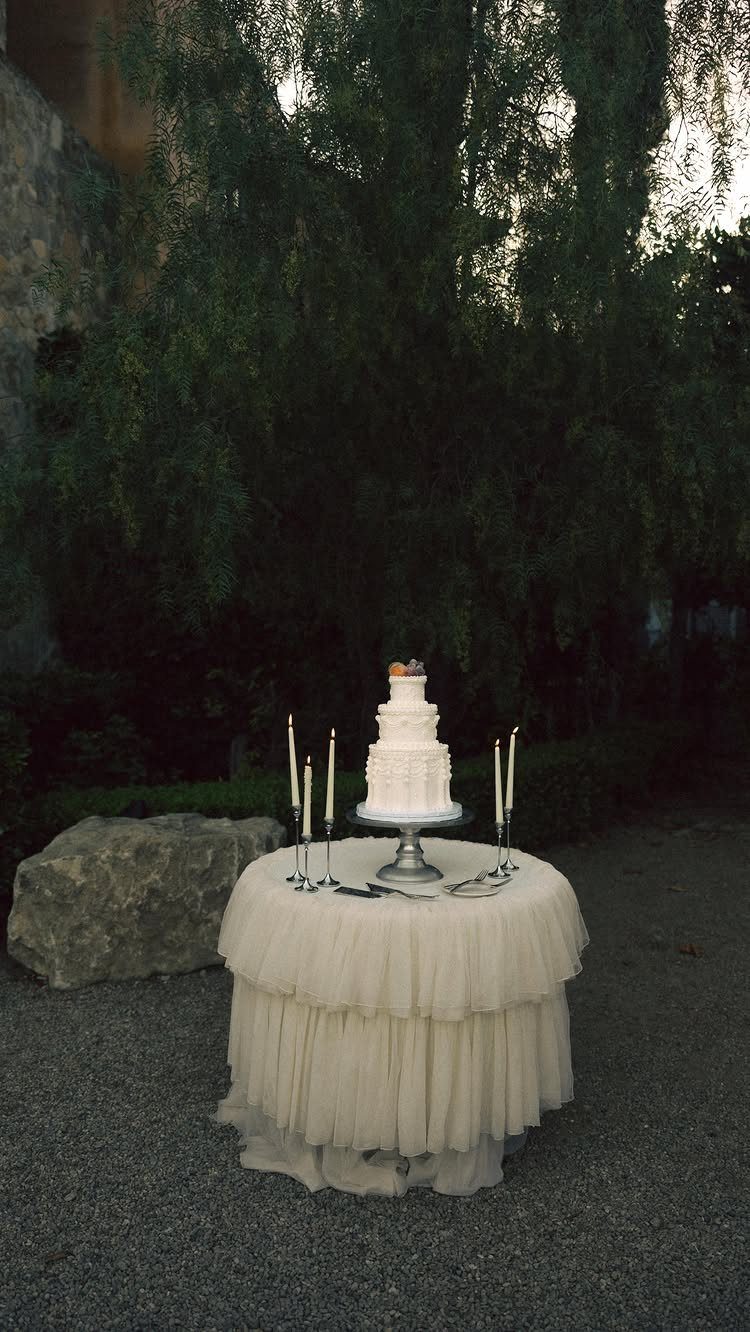 White wedding cake and taper candles on a ruffled cream tulle table outdoors.