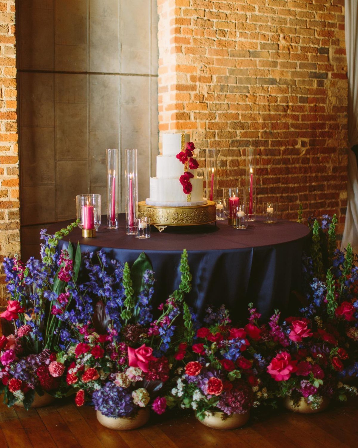 White wedding cake on gold stand surrounded by rich jewel-toned floral arrangements.