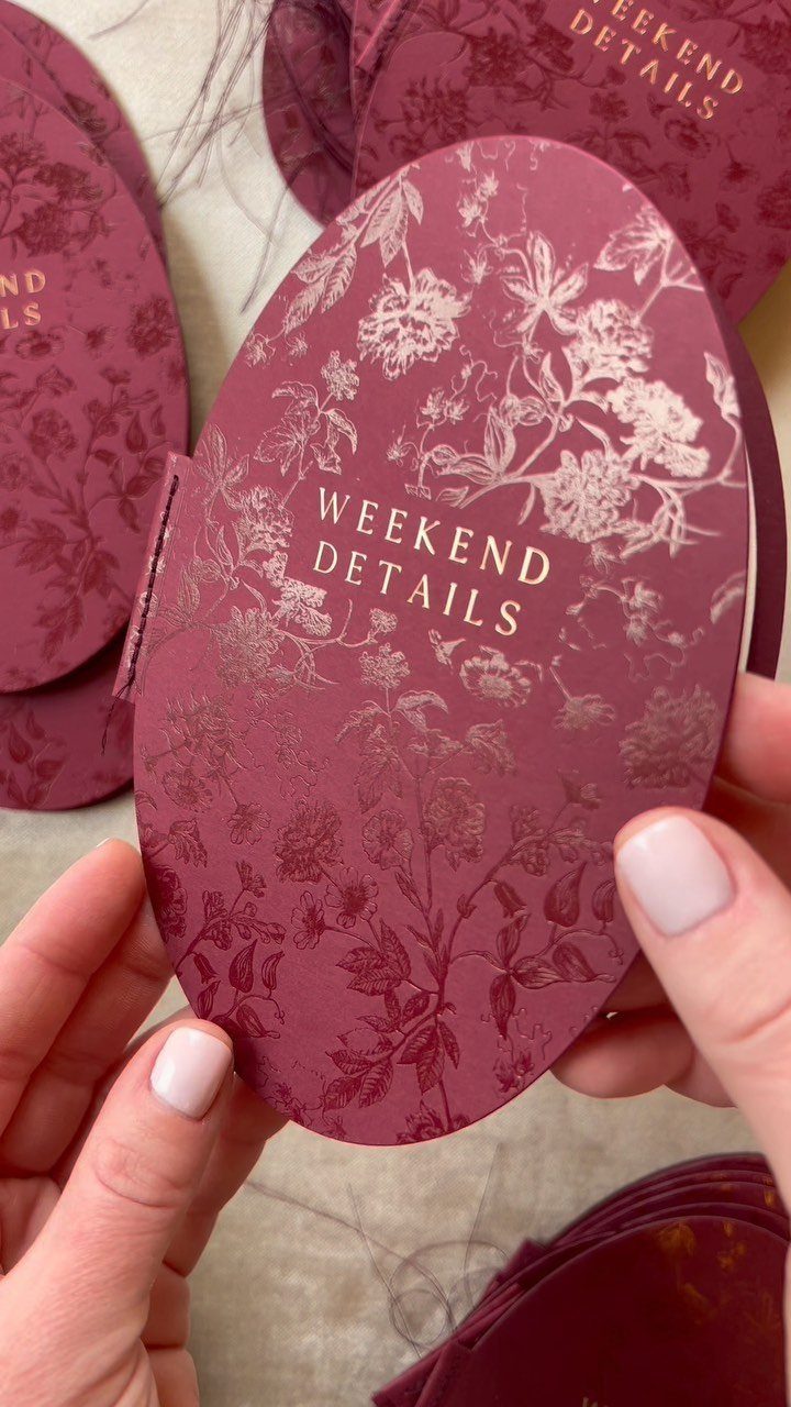 Burgundy oval wedding invitation card with gold floral patterns and gold text.