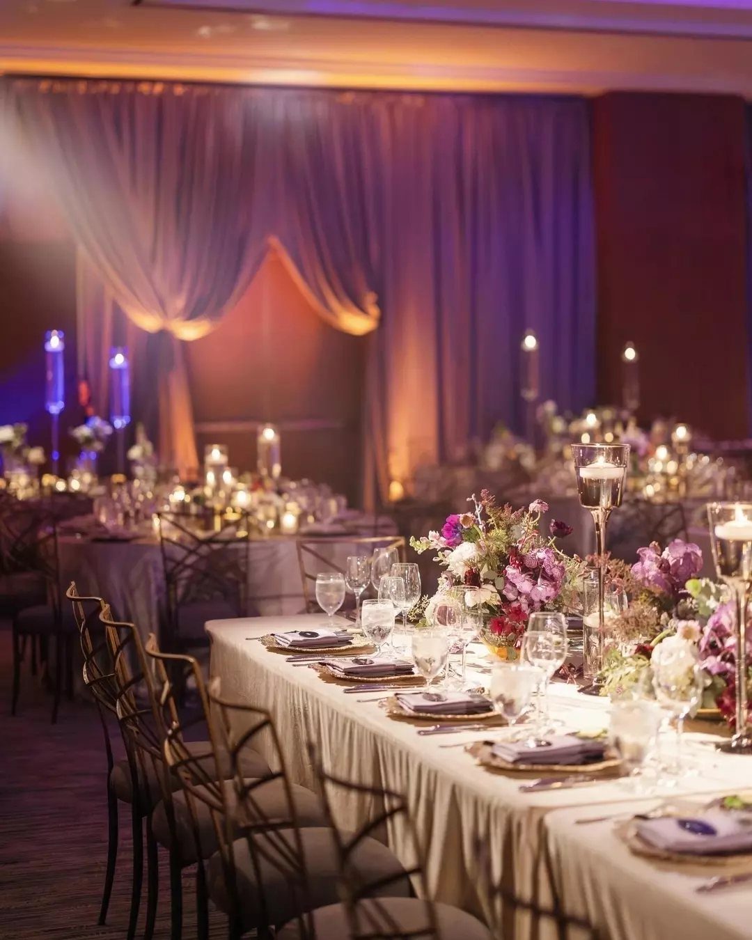 Wedding reception tables with purple flowers and warm amber uplighting against purple drapes.