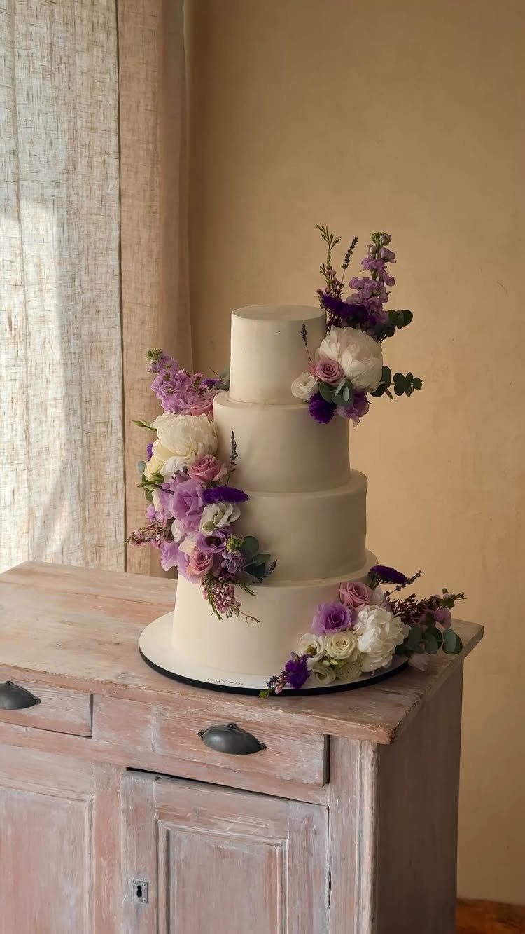 Four-tier cream wedding cake adorned with lavender and white floral arrangements on wooden furniture.