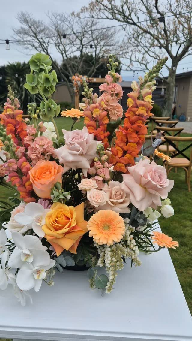 Peach and coral floral arrangement with roses and orchids on a white table.
