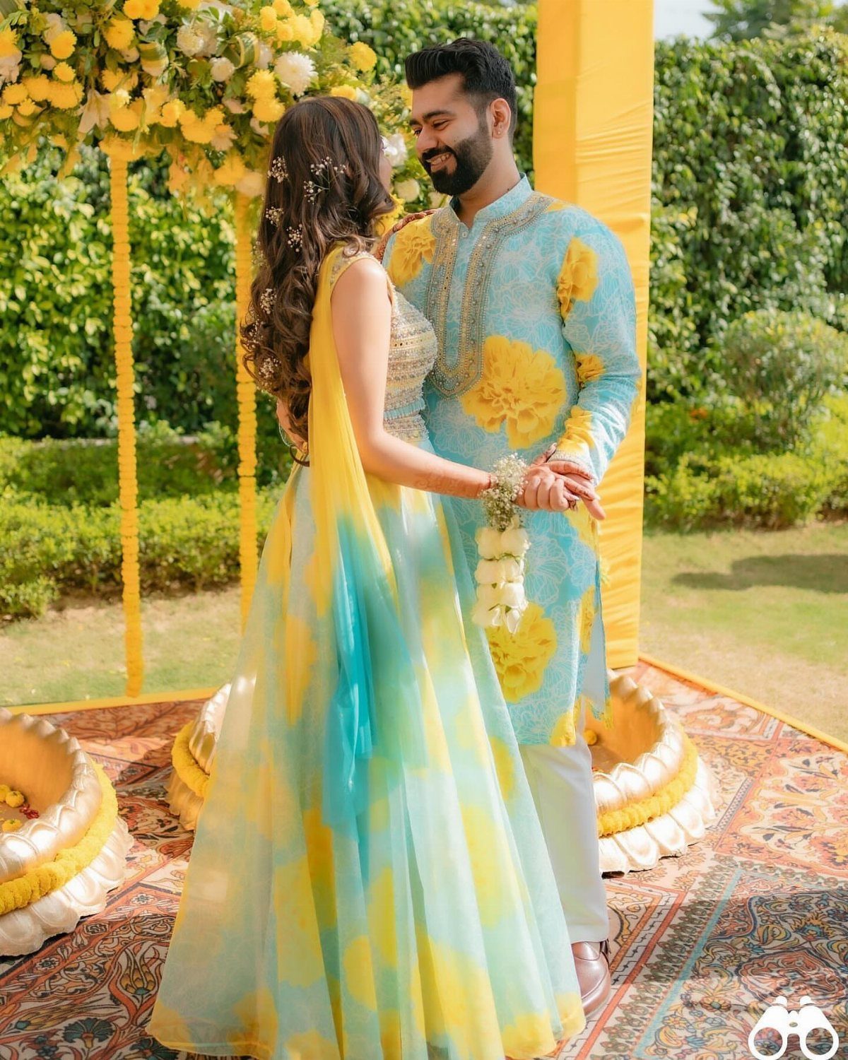 Couple in turquoise and yellow traditional Indian wedding attire standing amidst yellow flowers.