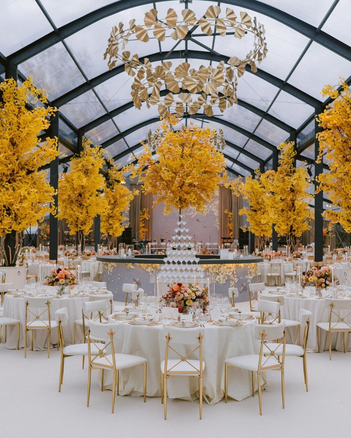 Luxury wedding reception with white tables, gold chairs, and large yellow floral arrangements.
