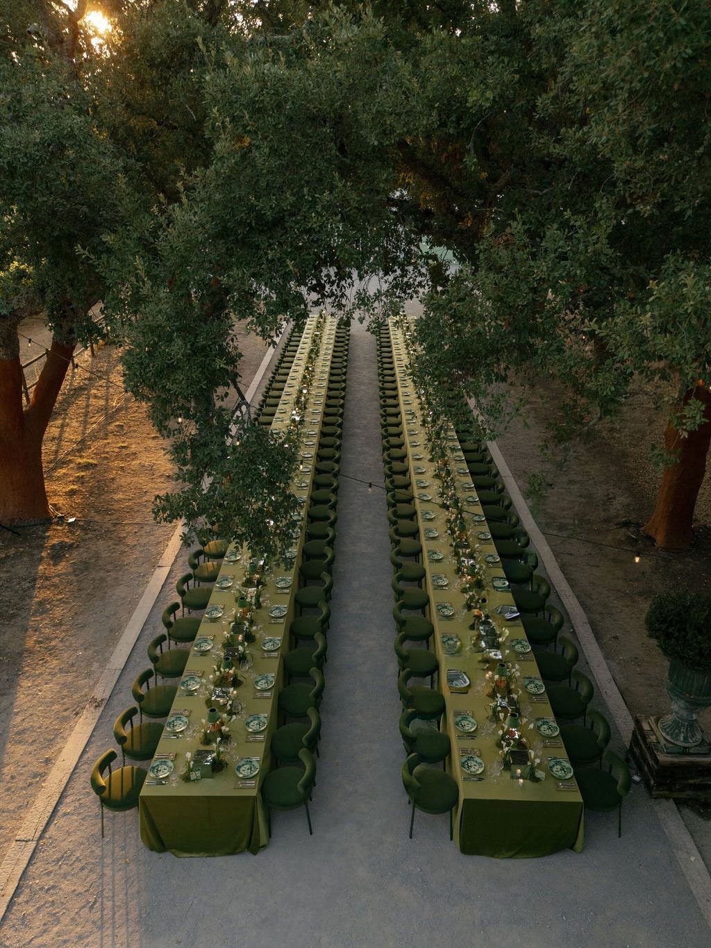 Two long dining tables with forest green linens set under large leafy trees.