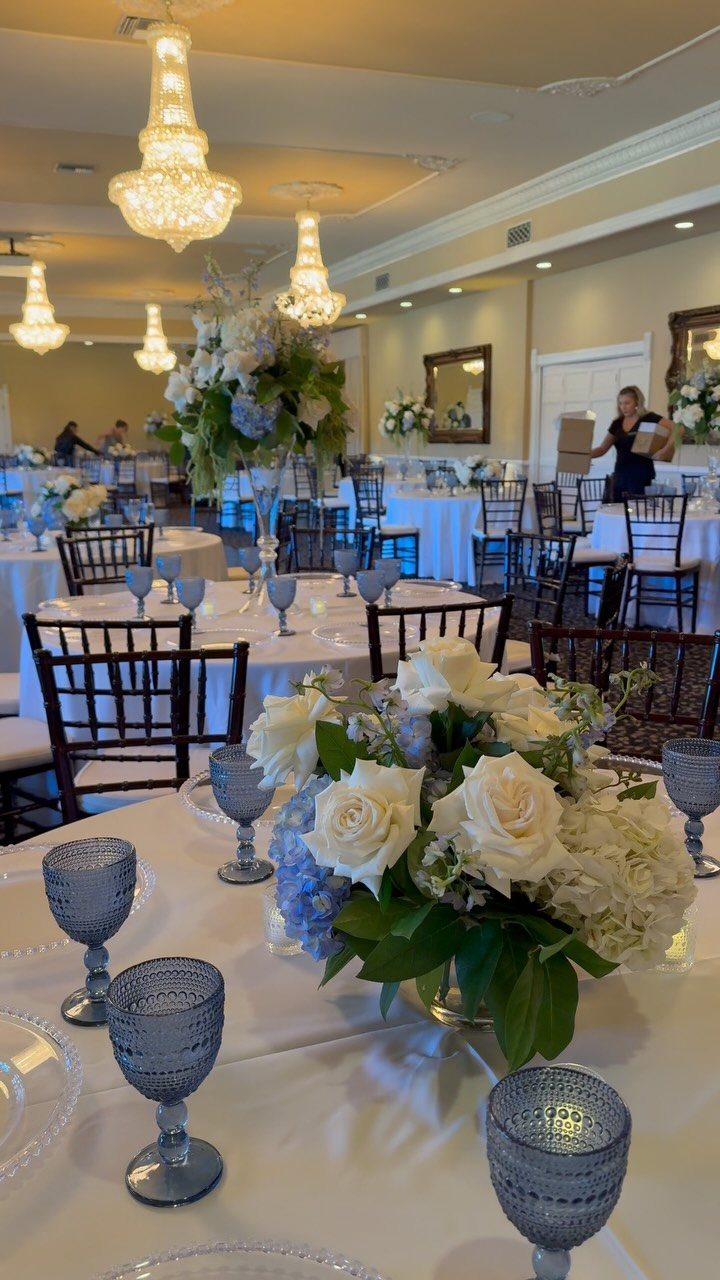 Wedding reception hall with white tables, light blue glassware, and white floral centerpieces.