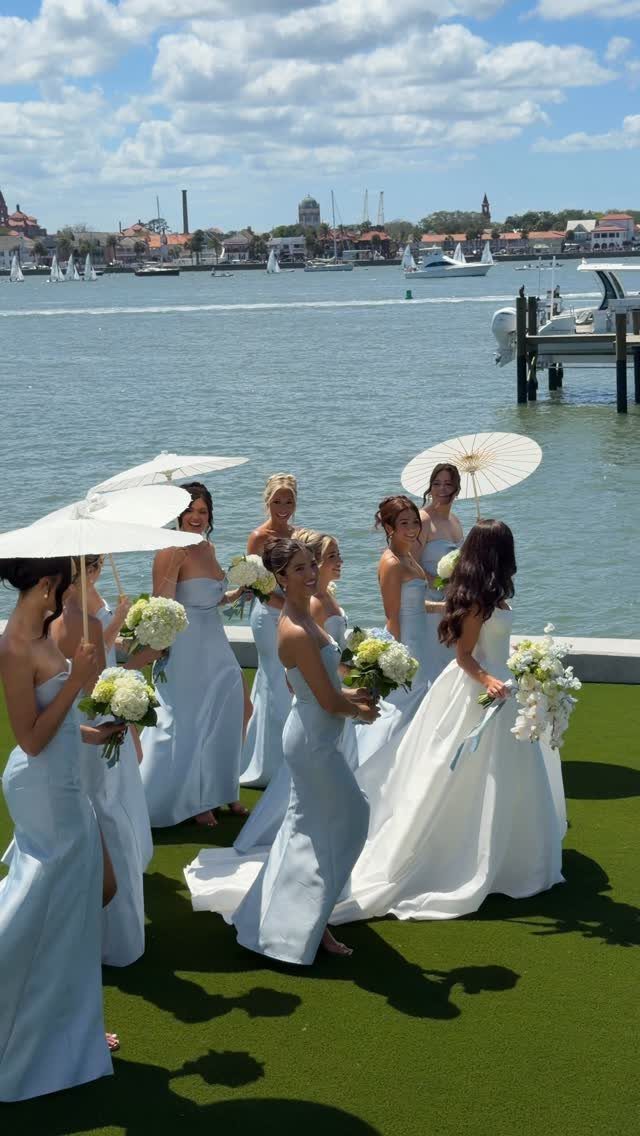 Bride in white and bridesmaids in light blue dresses by the waterfront.