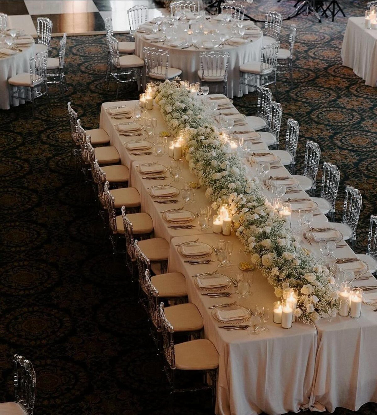 Long wedding head table with white floral runner, candles, and clear acrylic chairs.