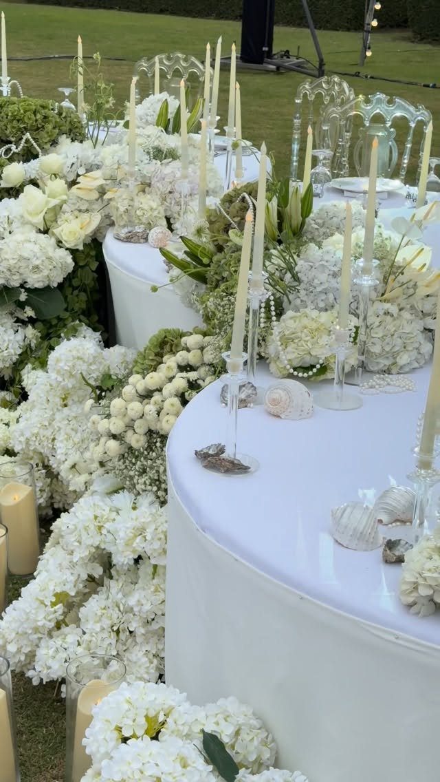 White round tables decorated with lush white flowers and tall white taper candles outdoors.