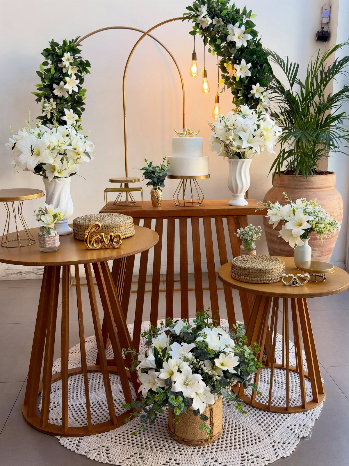 Wooden tables decorated with white flowers and a wedding cake under a gold arch.