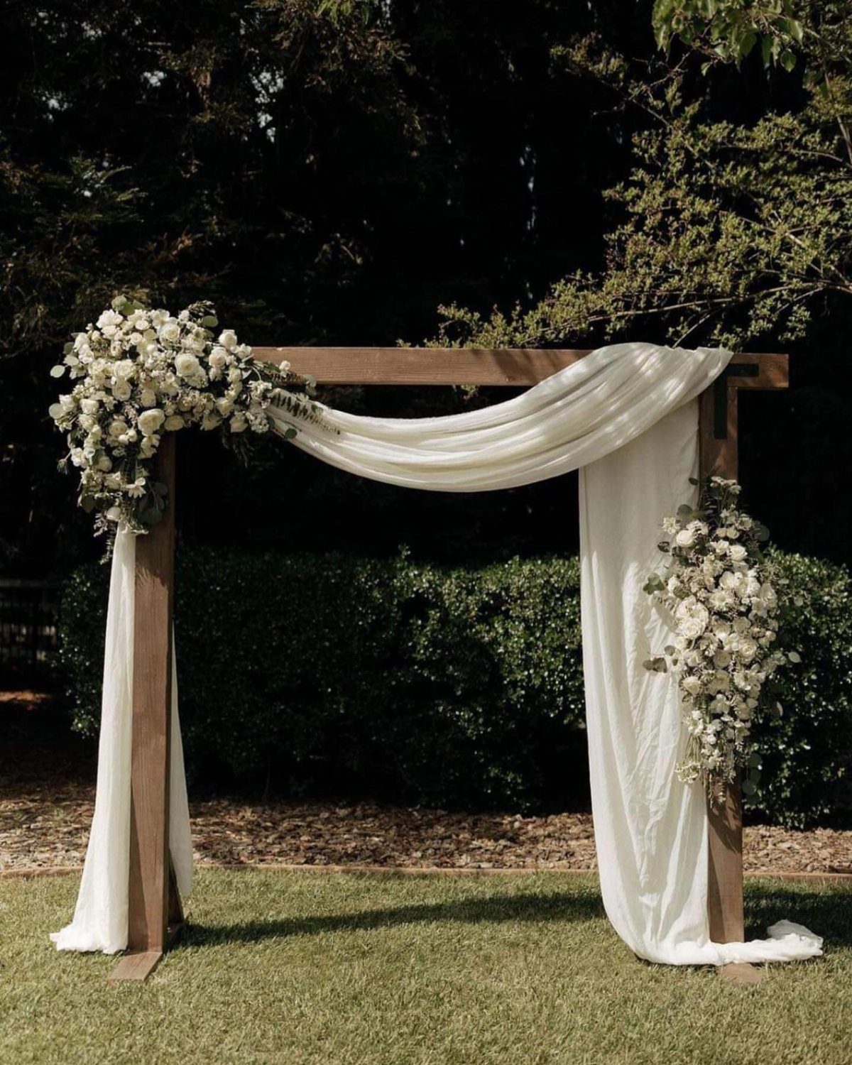 Wooden wedding arch draped in white fabric and decorated with white floral arrangements.
