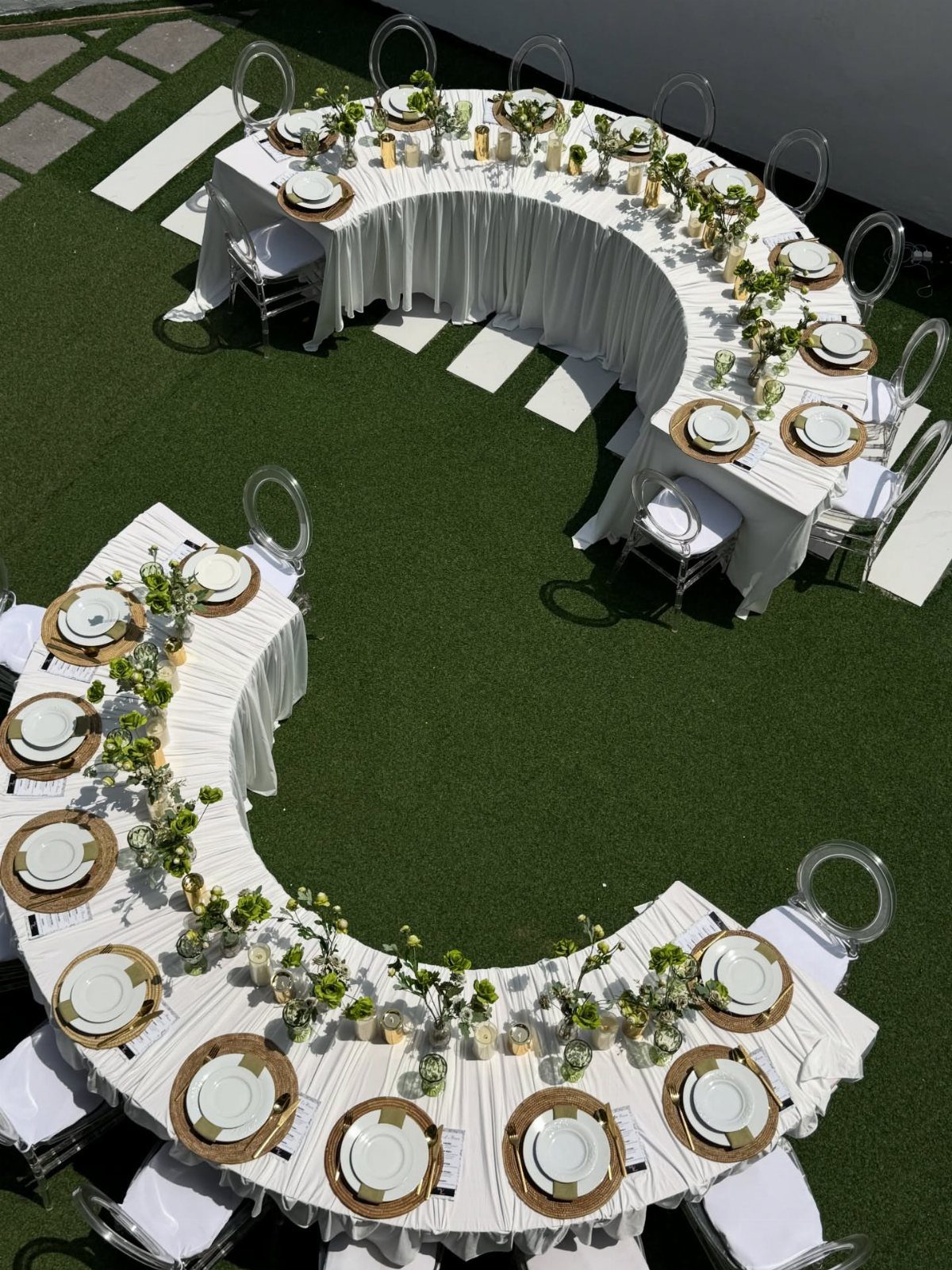 Curved white wedding tables with green foliage and clear chairs on green grass