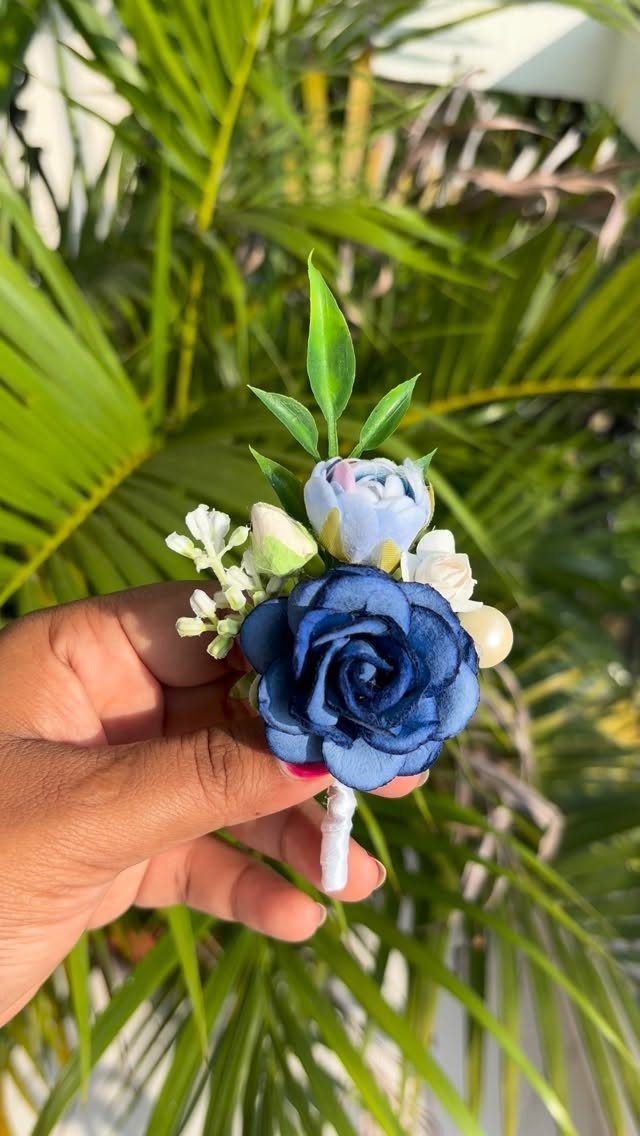 Navy blue rose boutonnière with light blue and white flowers held against palm leaves