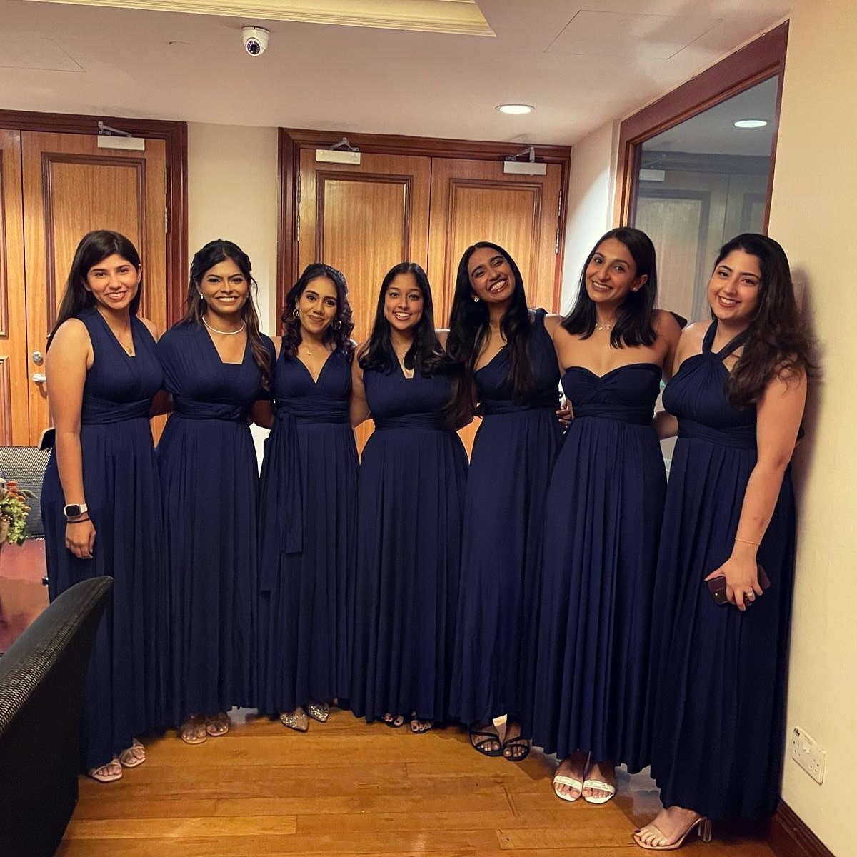 Seven women posing in long navy blue bridesmaid dresses in an indoor setting.