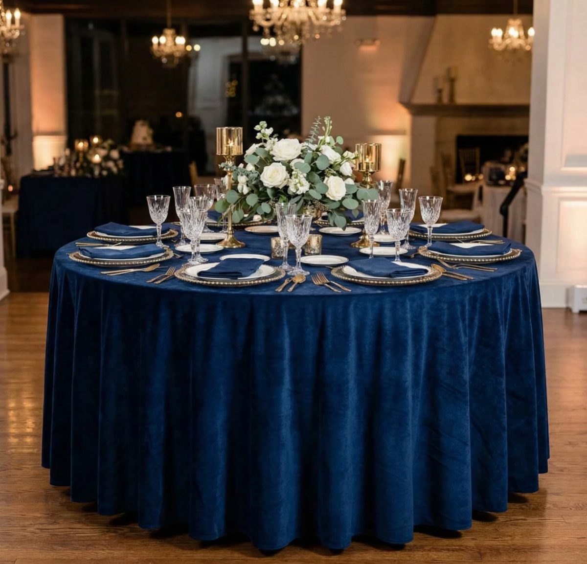 Round reception table with navy blue velvet tablecloth, gold chargers, and white flowers.