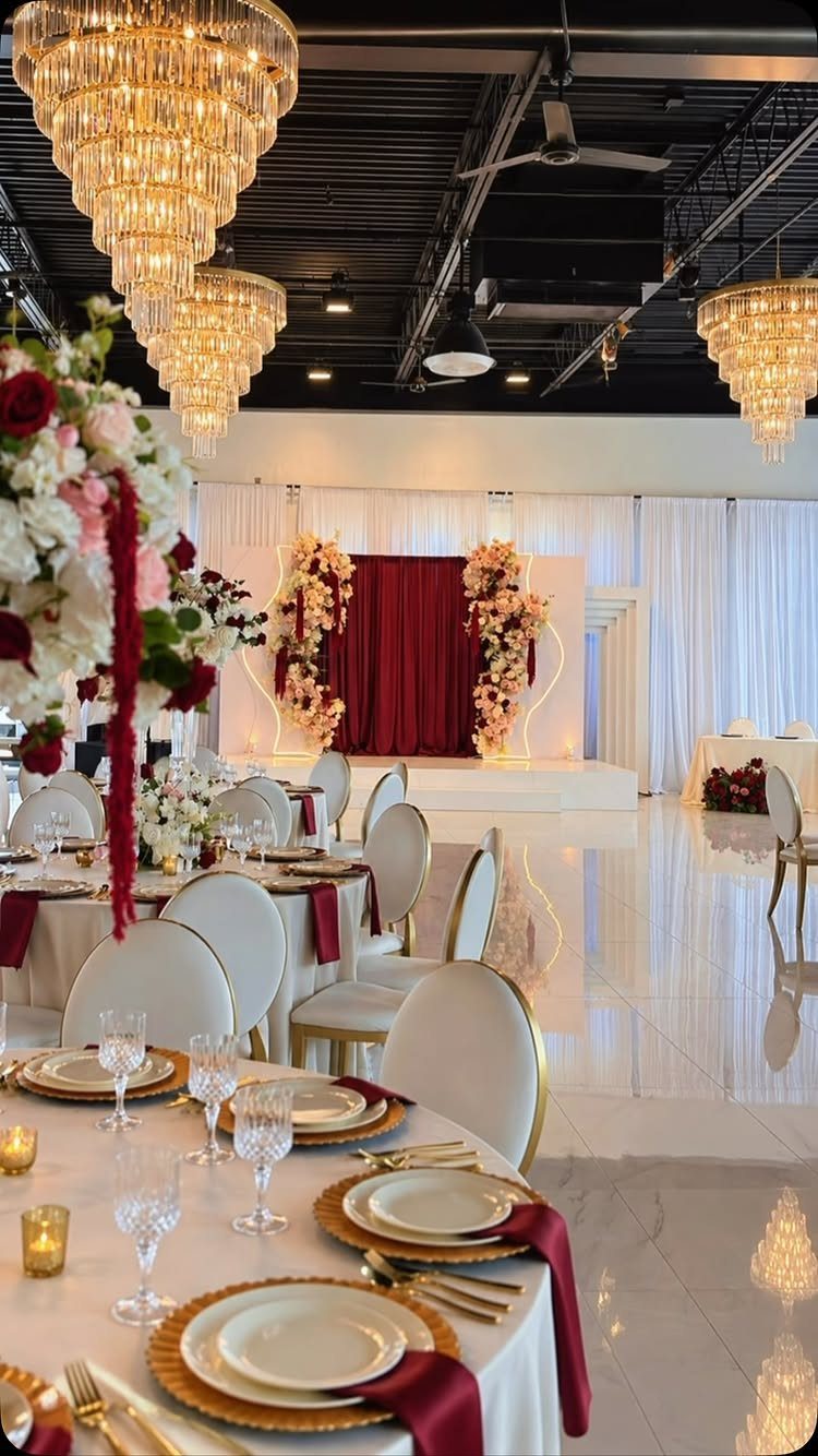 Wedding reception tables with burgundy napkins and gold plates under crystal chandeliers.