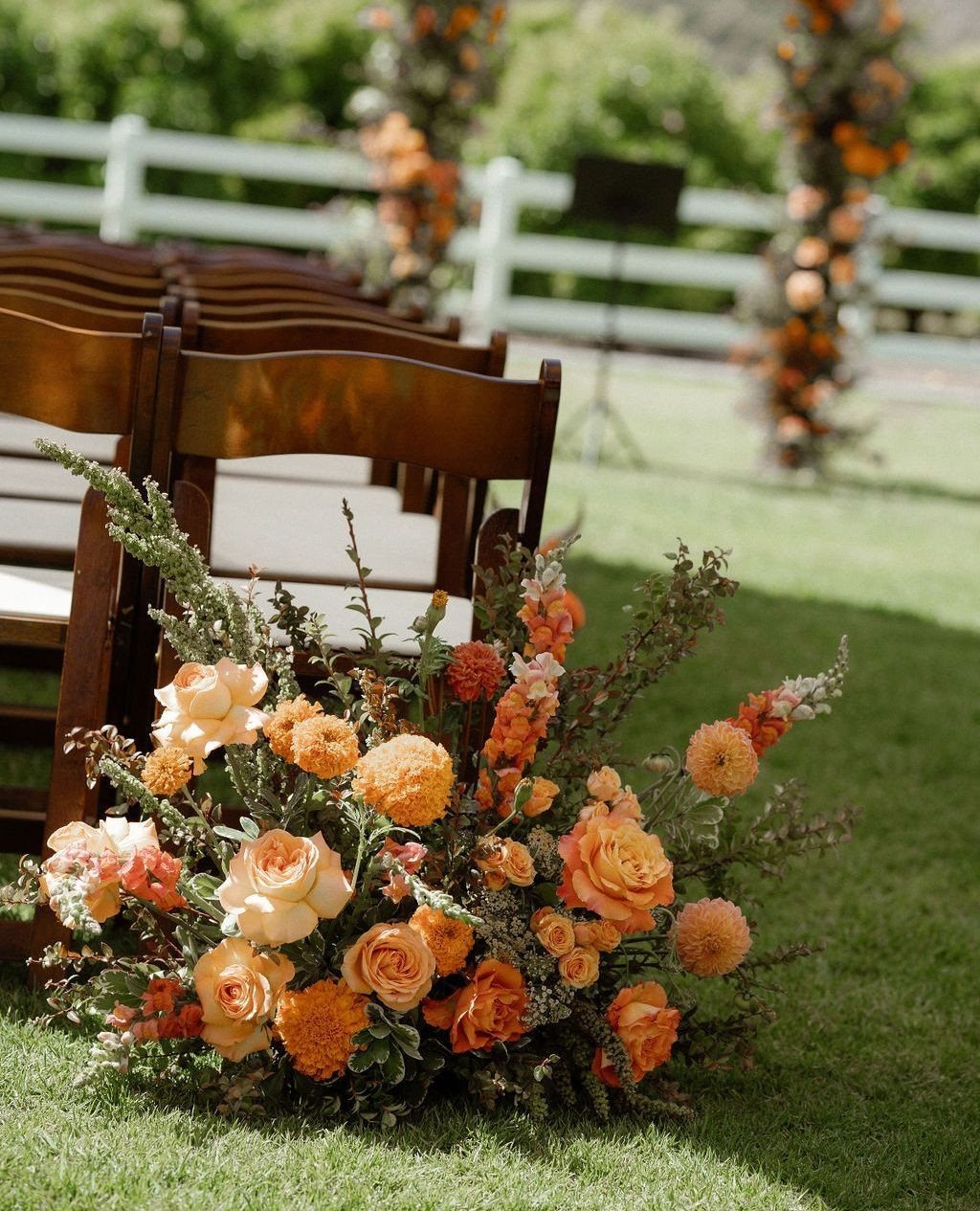 Muted orange floral arrangement with roses and greenery beside wooden wedding chairs.