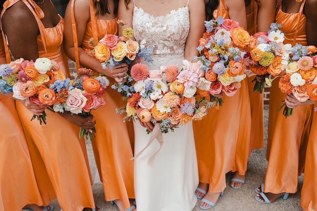 Bridesmaids in orange dresses holding bouquets of orange and blue flowers beside a bride.
