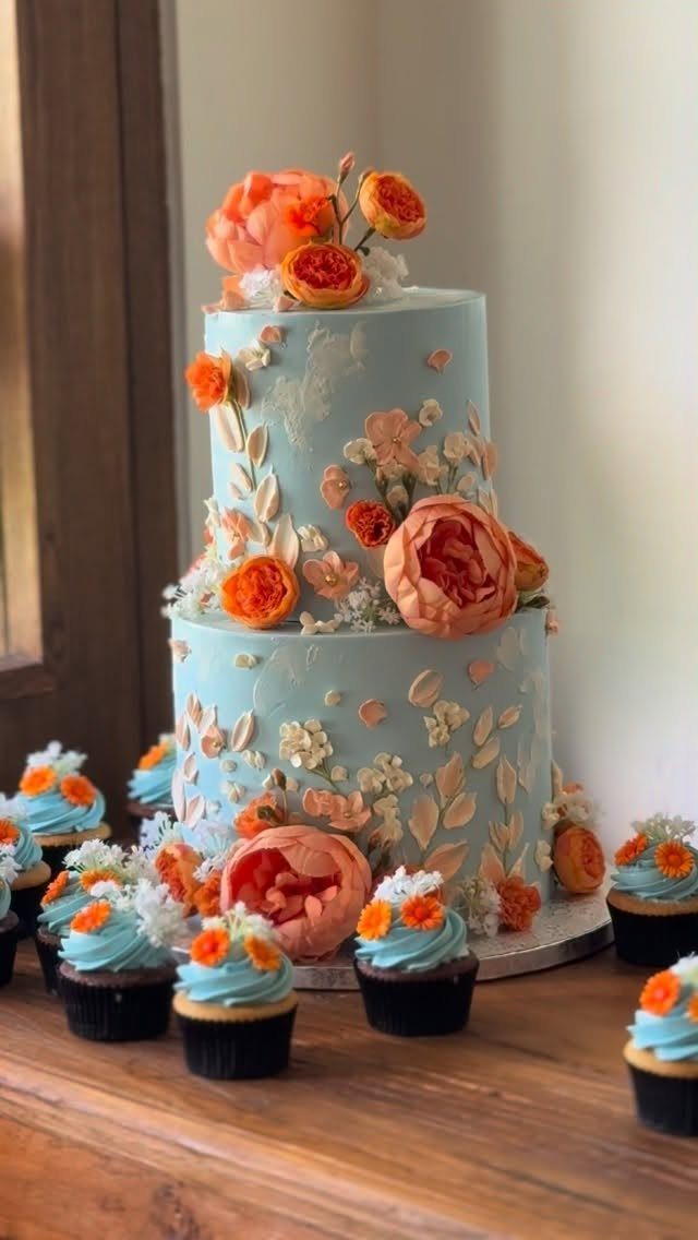 Two-tier light blue wedding cake with orange floral decorations and matching cupcakes.