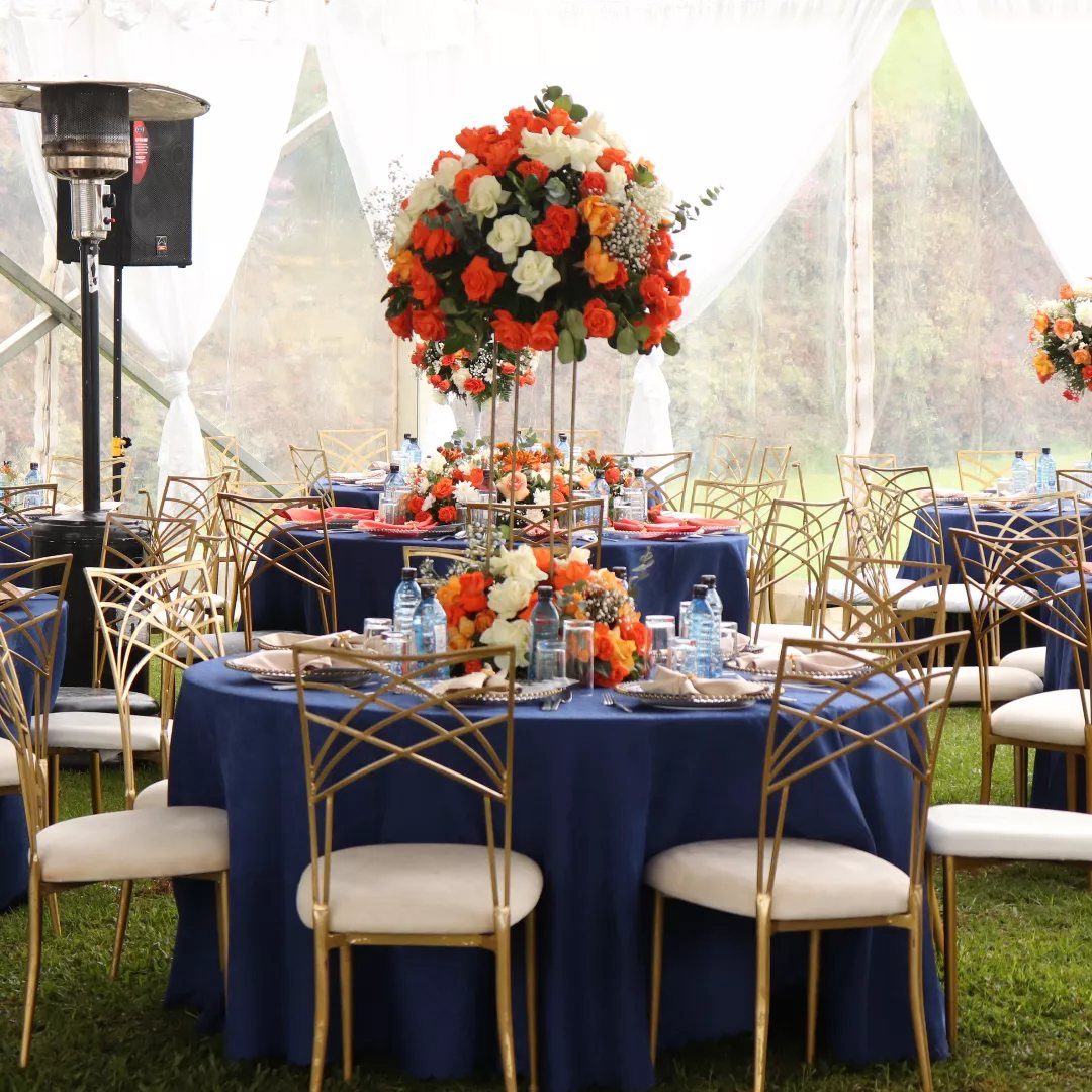 Navy blue tablecloths and gold chairs with orange and white floral centerpieces in a tent