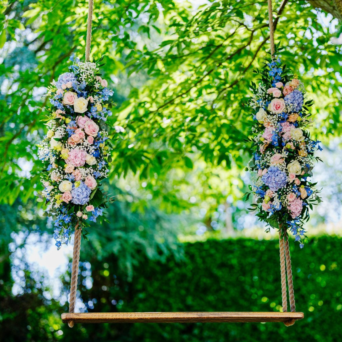 Wooden garden swing adorned with pink, blue, and white floral arrangements among green trees.