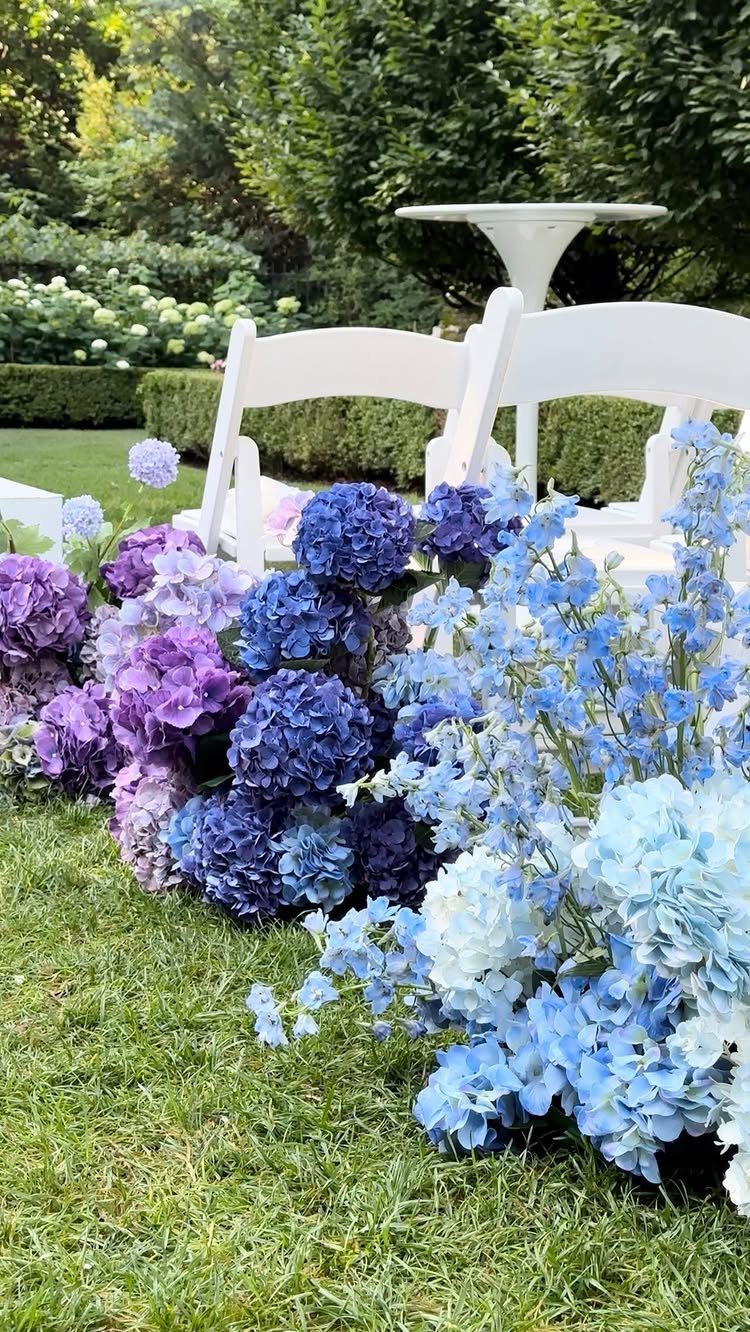 Purple and blue hydrangeas arranged on grass beside white wedding chairs in a garden.