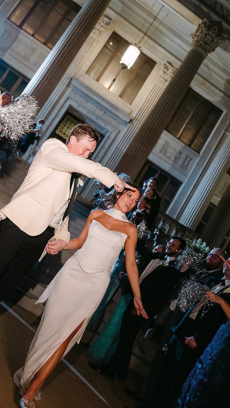 Bride and groom walking through a cheering crowd with silver pom-poms in a hall.