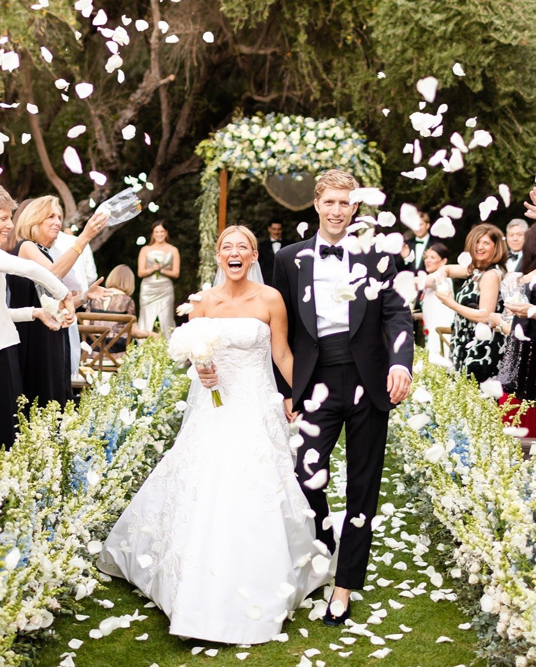 Bride and groom walking through a shower of white flower petals outdoors.