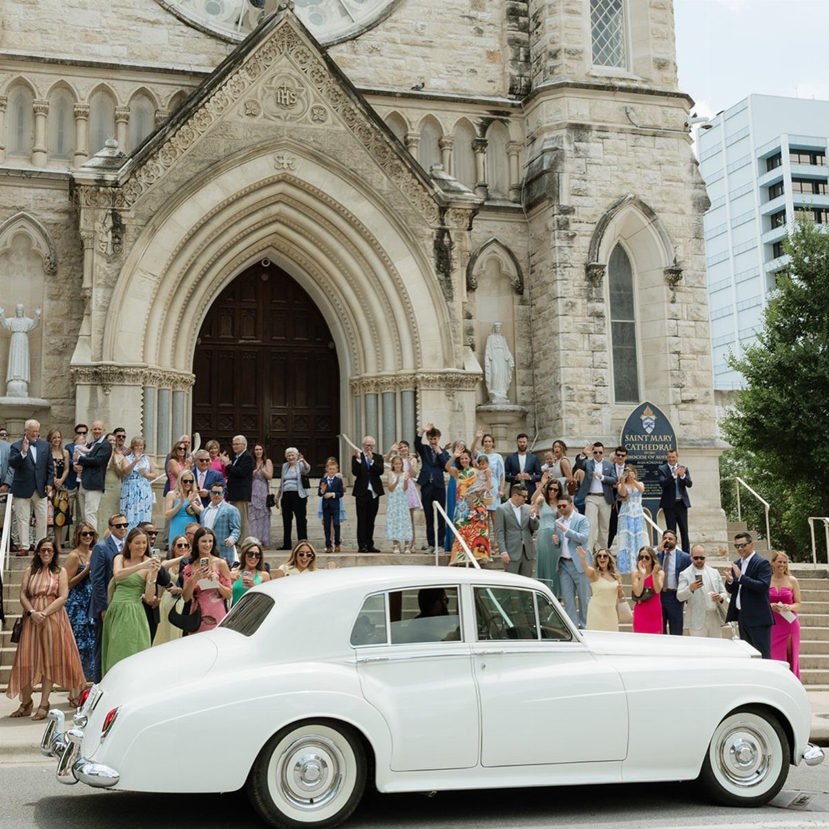 White vintage luxury car parked before a stone cathedral with cheering wedding guests.