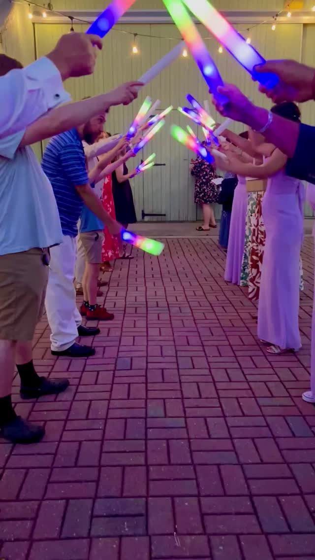 People holding colorful glowing neon sticks to create an archway on a brick path.