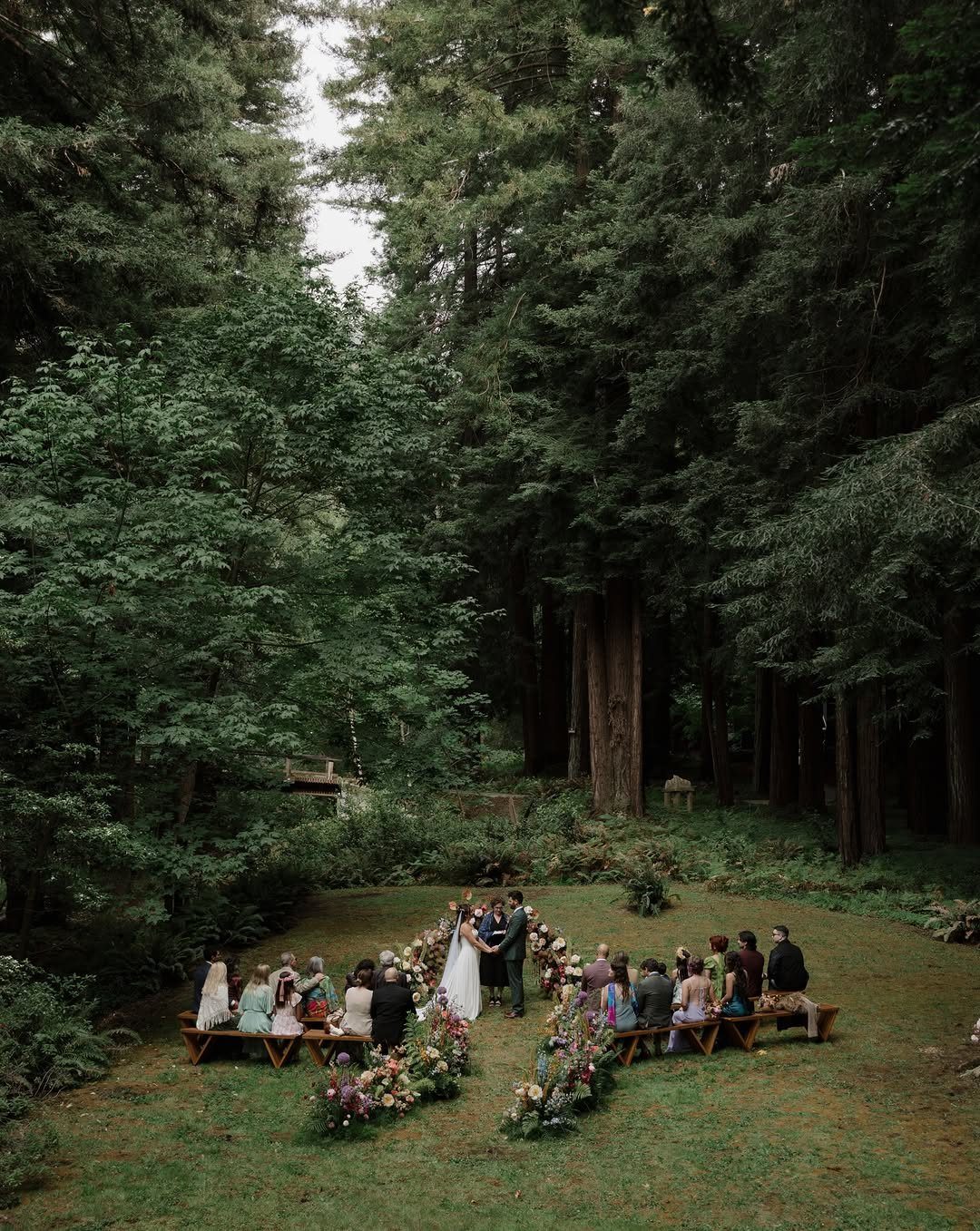Wedding ceremony in a forest with guests on wooden benches and floral decor.