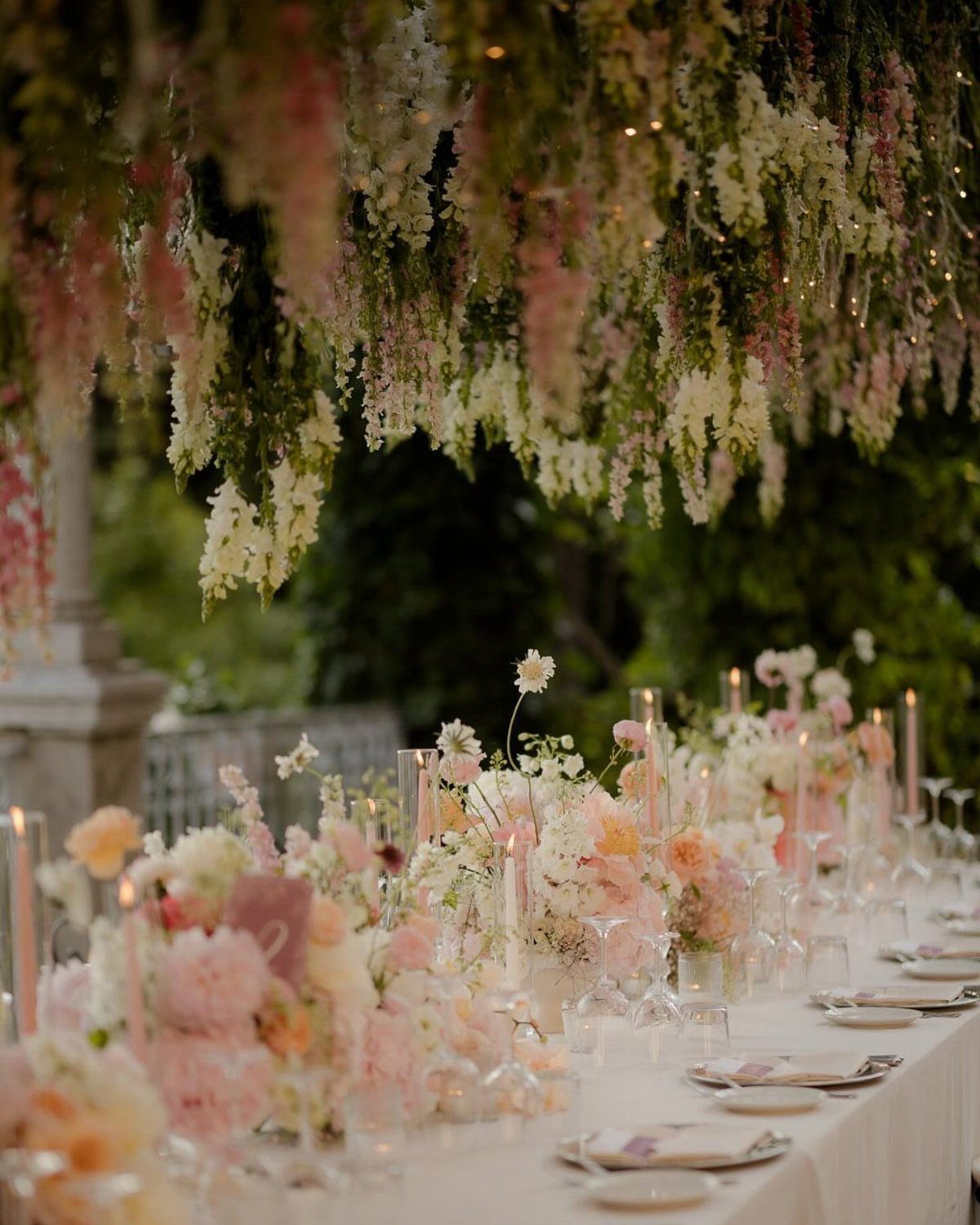 Floral arrangements and candles on a white table under a hanging flower canopy.