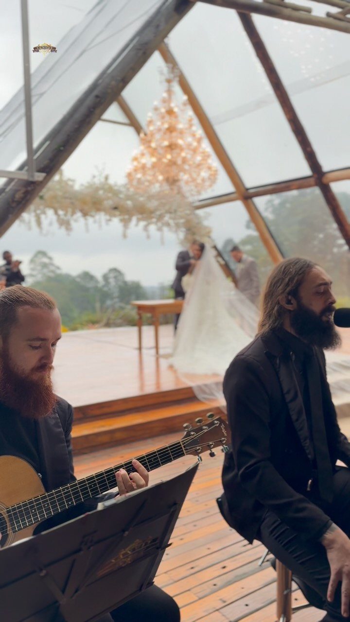 Musicians playing guitar at a wedding ceremony inside a glass pavilion with a chandelier.