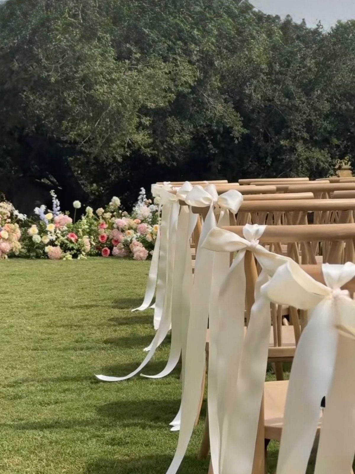 Cream satin bows tied to wooden chairs on a lawn with floral backgrounds.