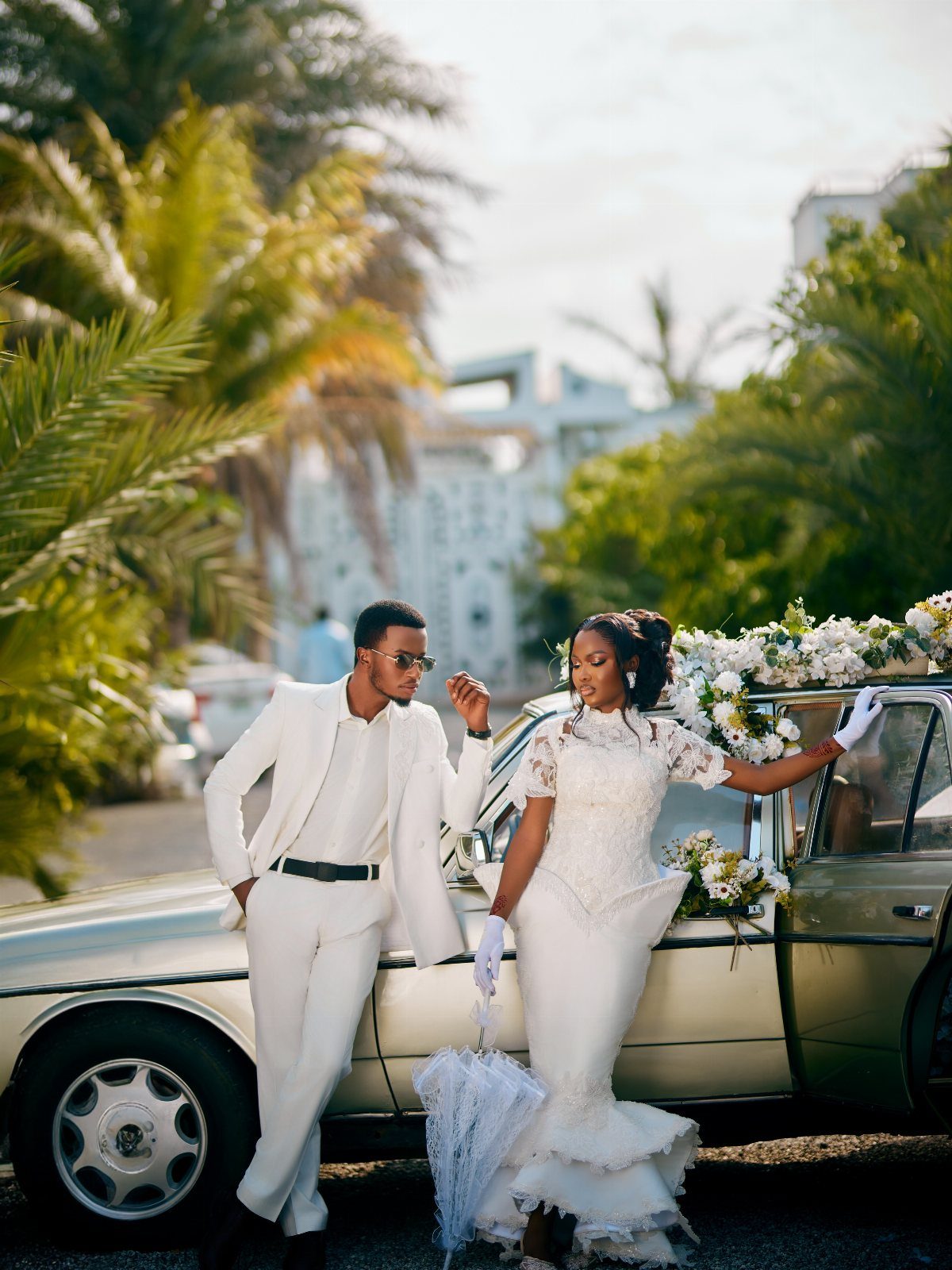 Wedding couple in white attire posing beside a vintage gold car with floral accents